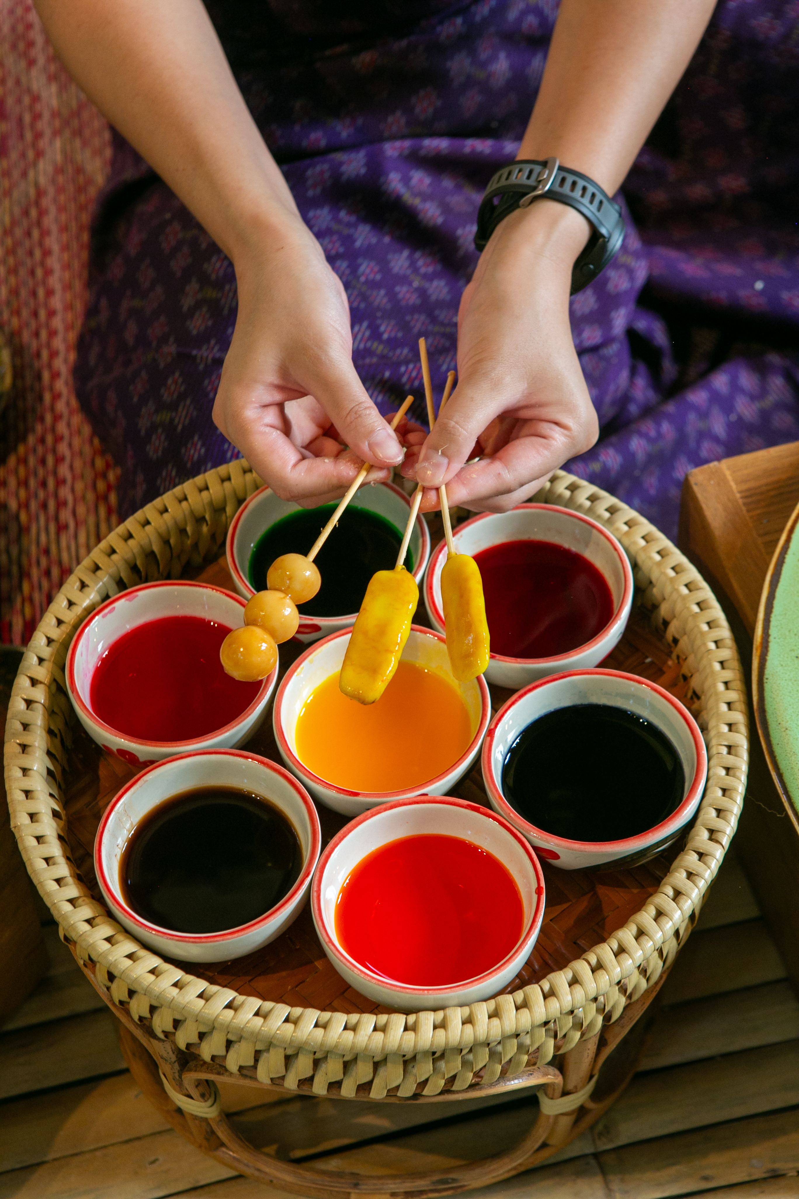 Hands dipping skewered Thai lookchoup desserts into a wheel of colorful dye bowls at a Poomjai Garden workshop.