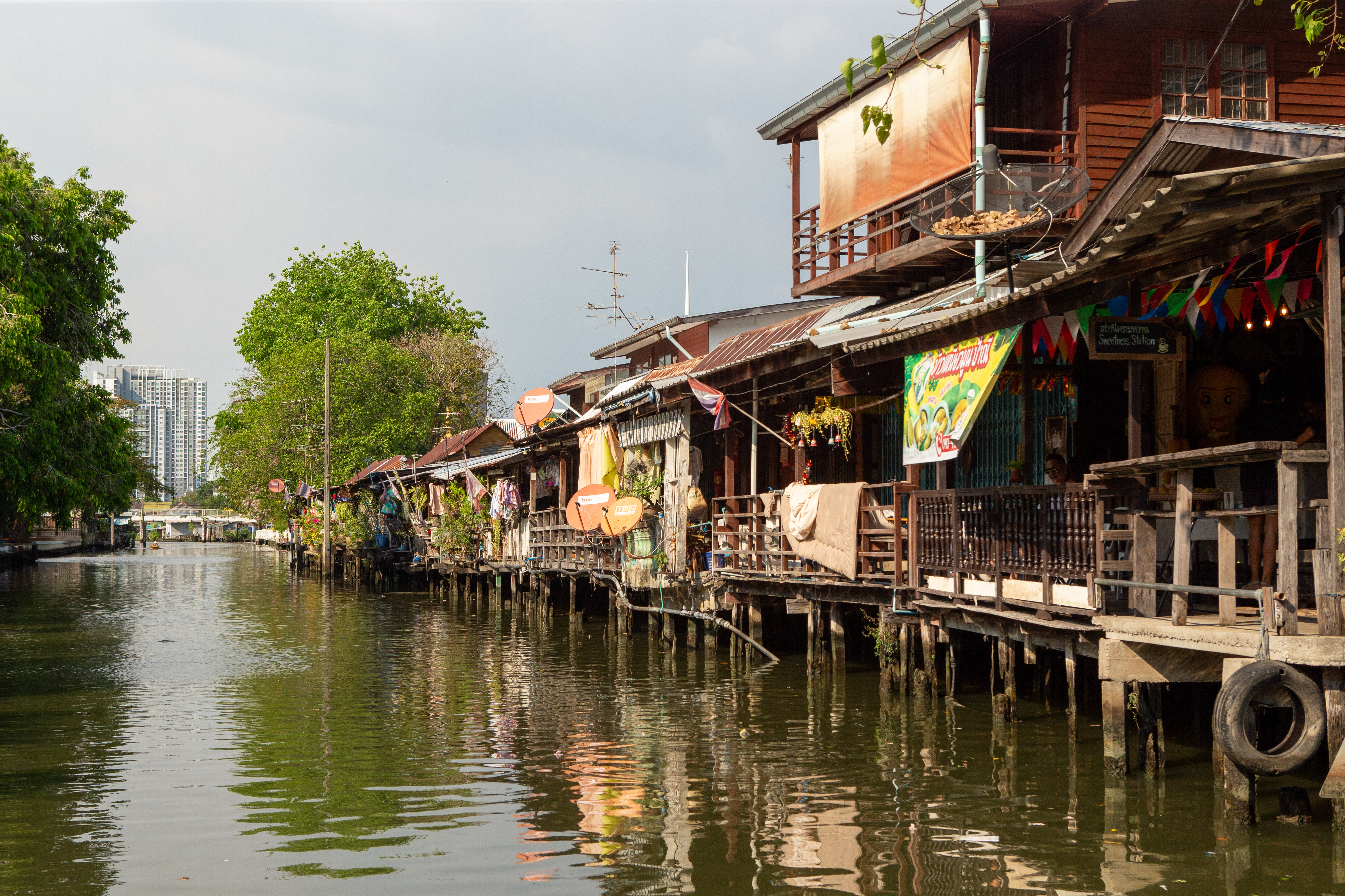 Canal-side stilt houses along the Thonburi waterway seen from the boat.