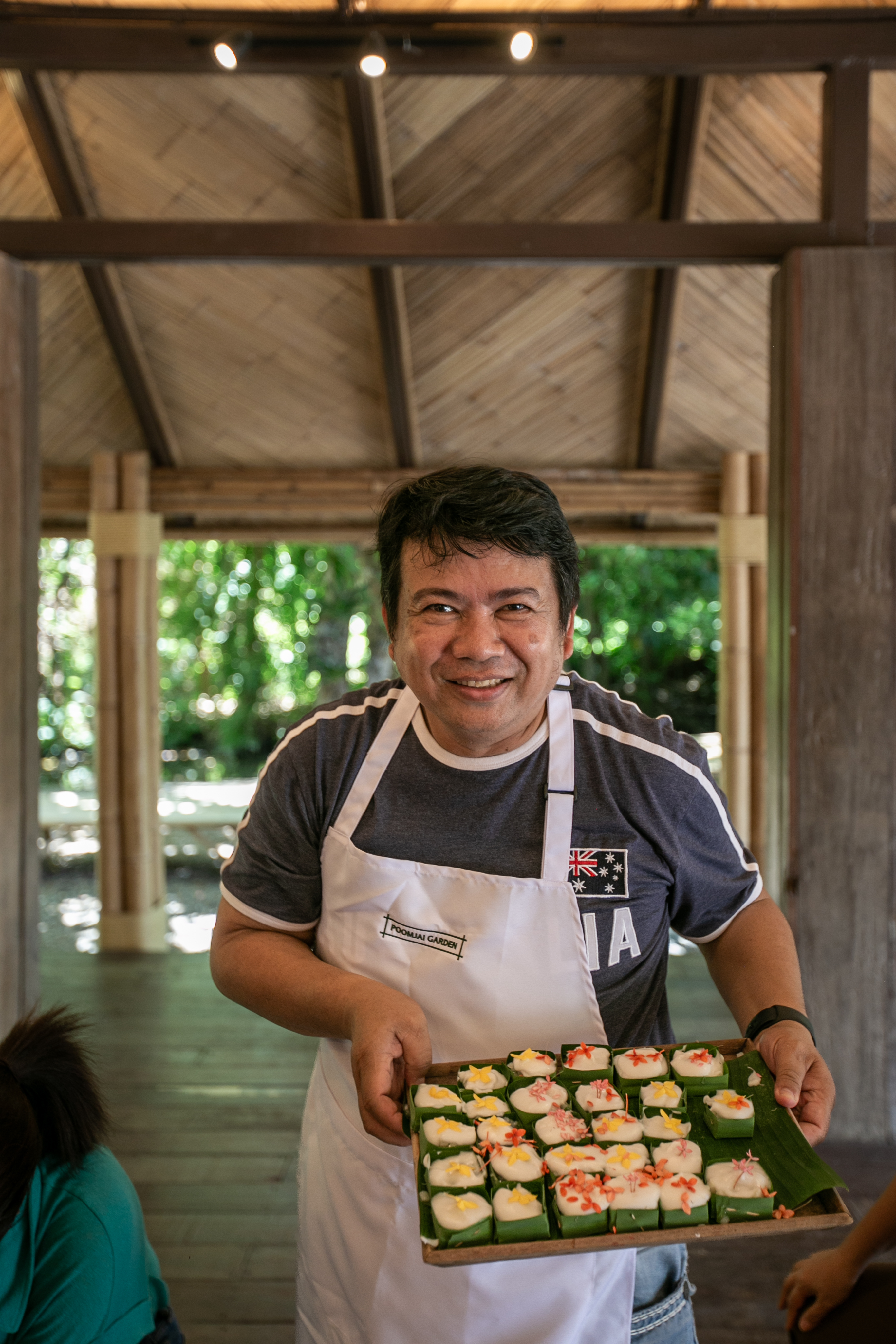 A host presenting a tray of finished Thai desserts wrapped in banana leaf.