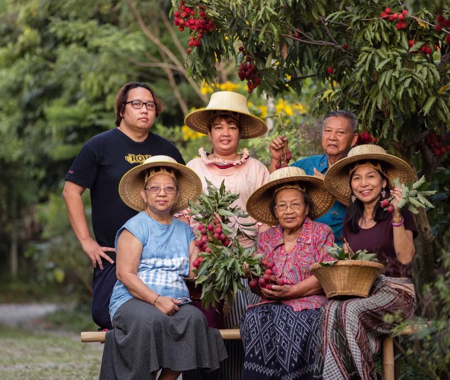 The family behind Poomjai Garden today, wearing traditional hats and holding freshly picked lychee branches under an orchard tree.