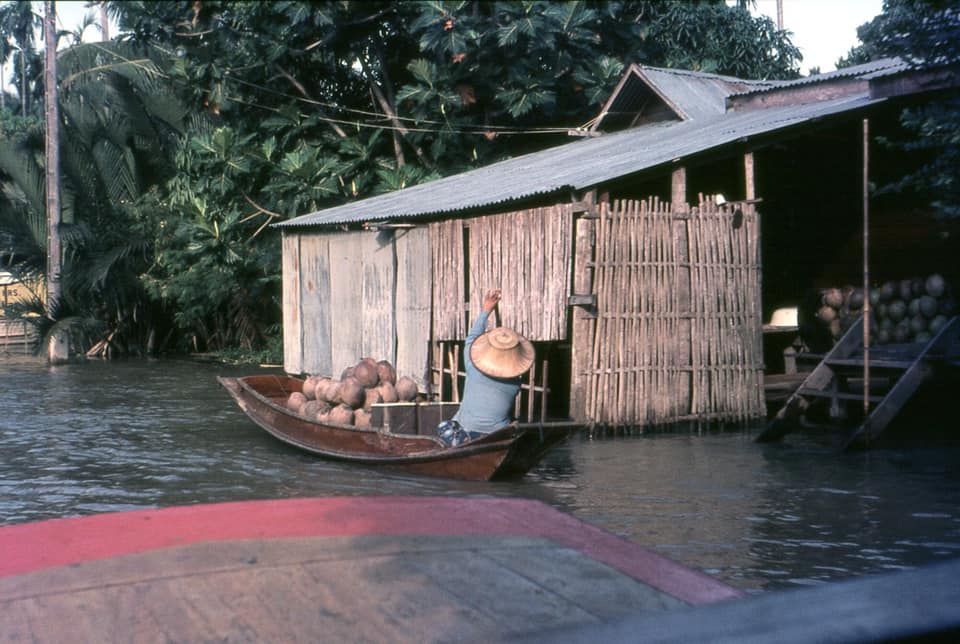 Archival photo of a wooden house on stilts over the canal, with a man rowing a small boat toward it.