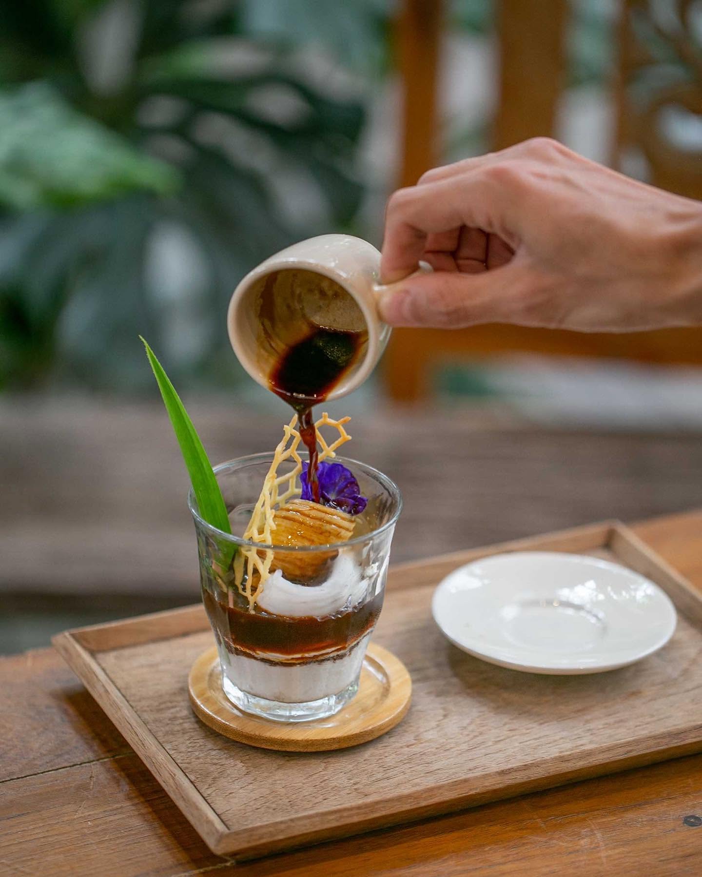 A coffee sauce being poured over a layered dessert with a pandan leaf garnish at Natura Caf&eacute;.